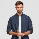 waist-up-portrait-handsome-serious-unshaven-male-keeps-hands-together-dressed-dark-blue-shirt-has-talk-with-interlocutor-stands-against-white-wall-self-confident-man-freelancer_273609-16320.jpg